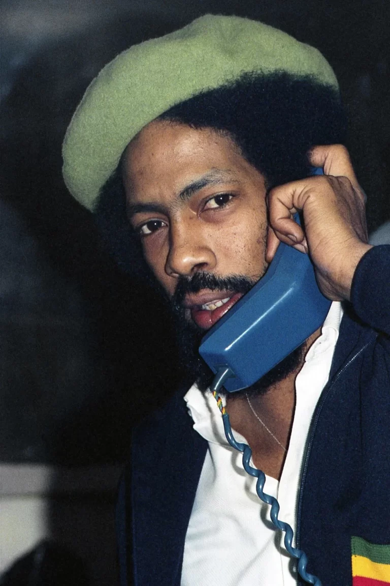 Jackie Mittoo sporting a green beret, holds a blue telephone handset with a captivating expression for a photo shoot.