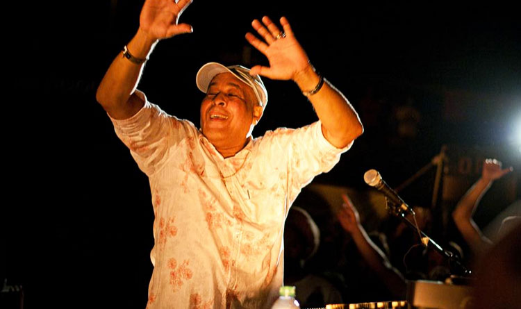 King Jammy with his arms raised in motion, wearing a beige baseball cap and blouse