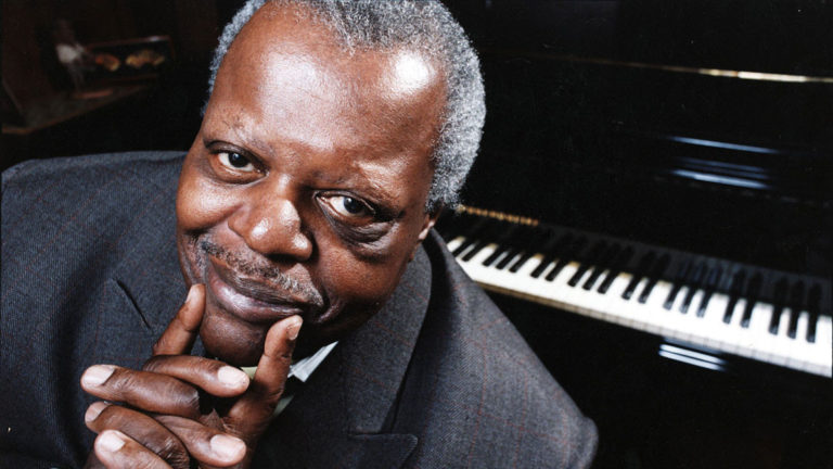 Portrait of Oscar Peterson wearing a gray blazer with a piano in the background