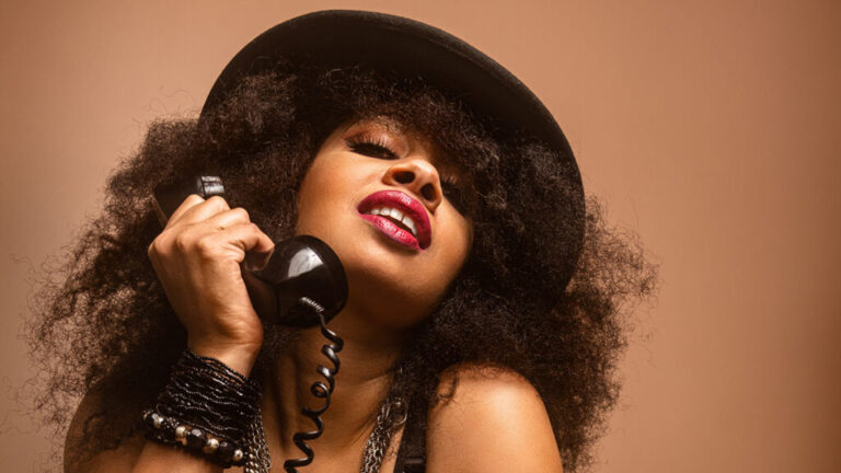 Melanie Durrant holding black handset of rotary phone and wearing a hat against a brown background for a captivating photo shoot.