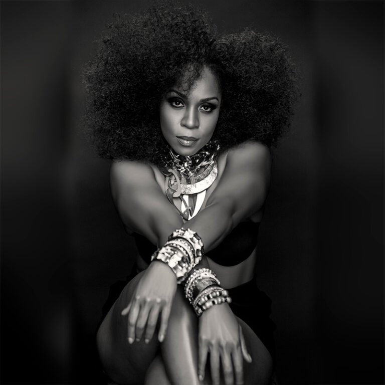 Striking black and white of Divine Brown adorned with jewelry and her arms crossed, showcasing her elegance and style.