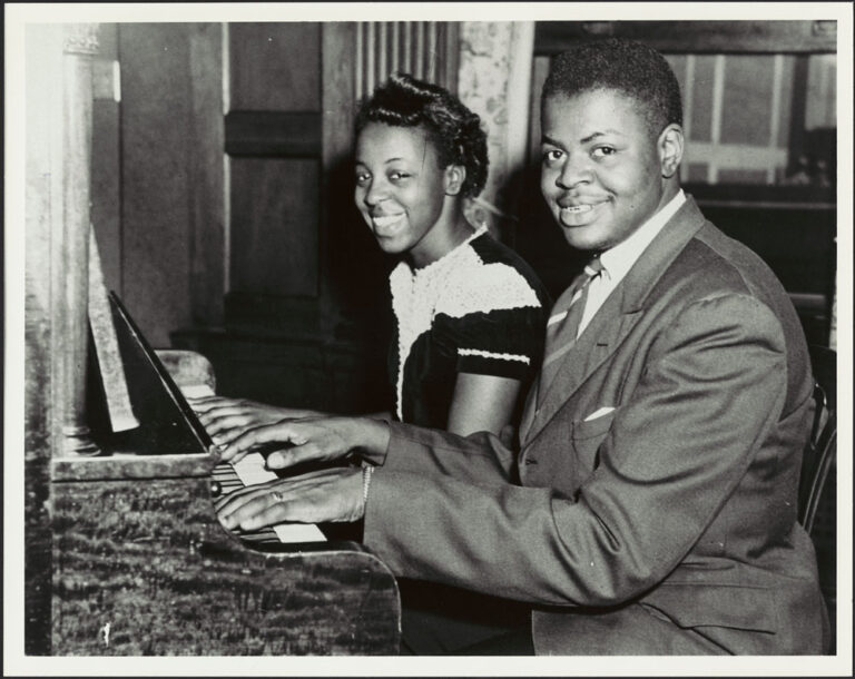 Daisy Peterson Sweeney and her brother Oscar Peterson looking at the camera while playing the piano, capturing a moment of connection among the Peterson family.