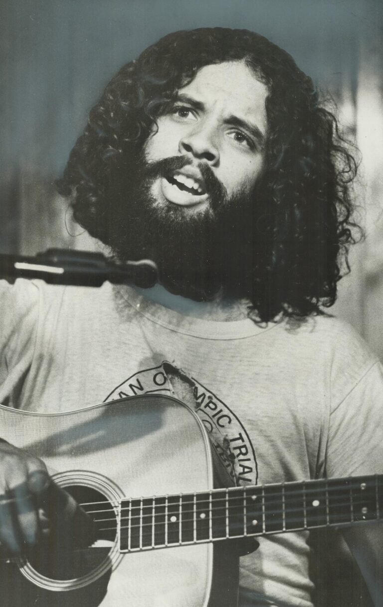 Dan Hill singing into microphone while playing the guitar at his performance.