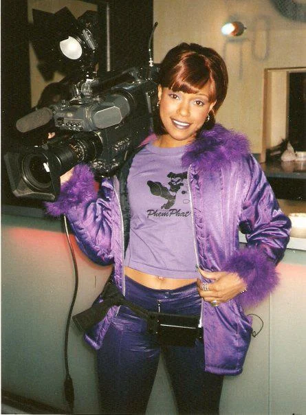 Michie Mee in purple fur jacket and purple garments, smiling and holding a broadcasting camera