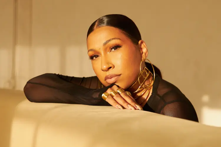 Melanie Fiona dressed in black and wearing gold jewelry, resting on couch and looking into camera