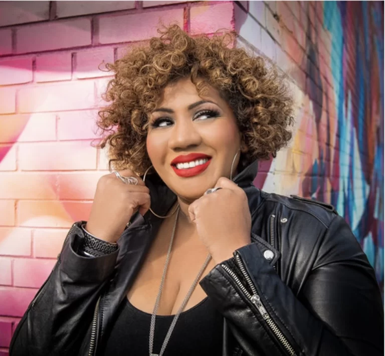 Simone Denny in front of brick graffiti wall, collar upturned and smiling, looking off to side