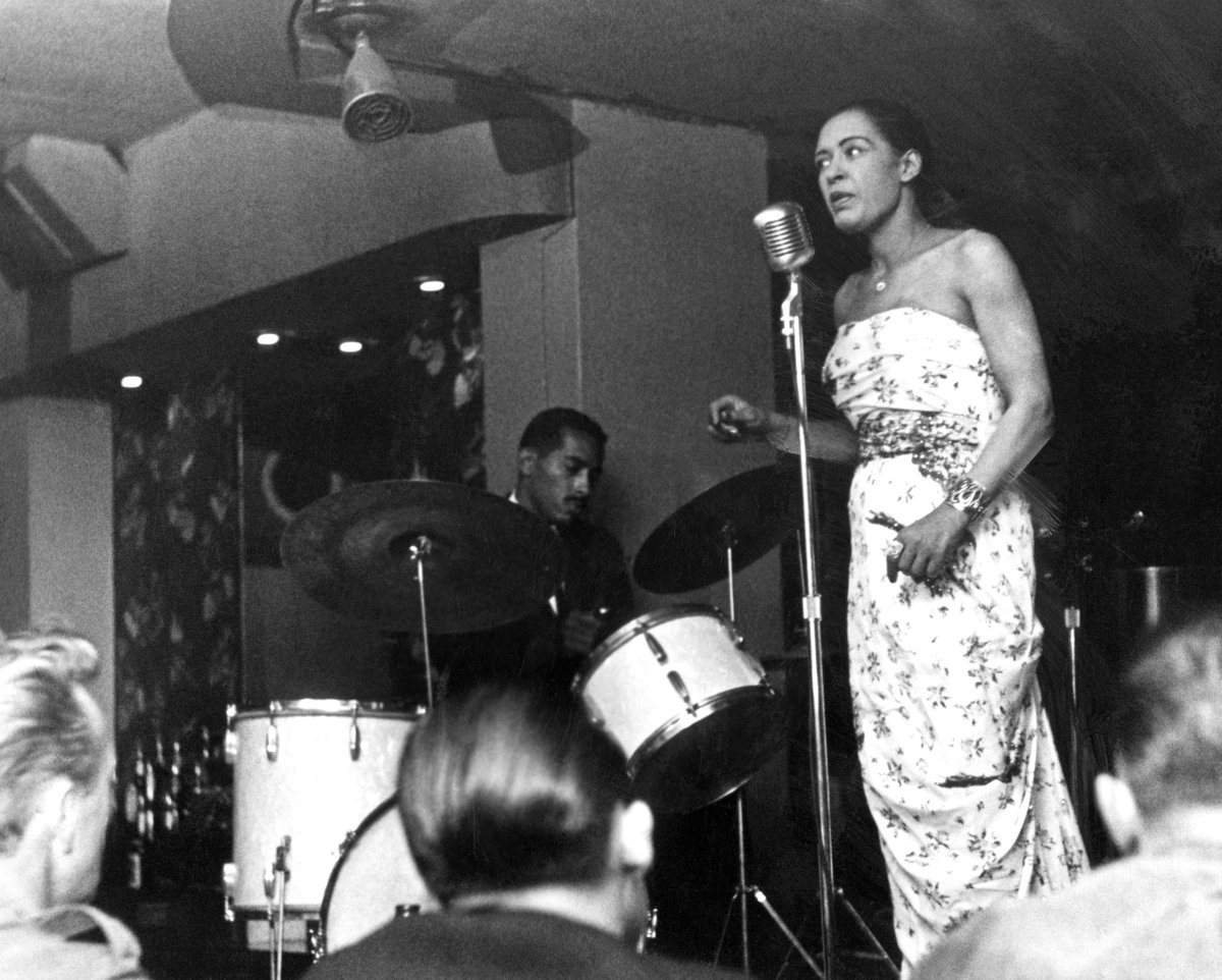 black and white photograph of an artist singing on stage with a crowd, and drummer in the background