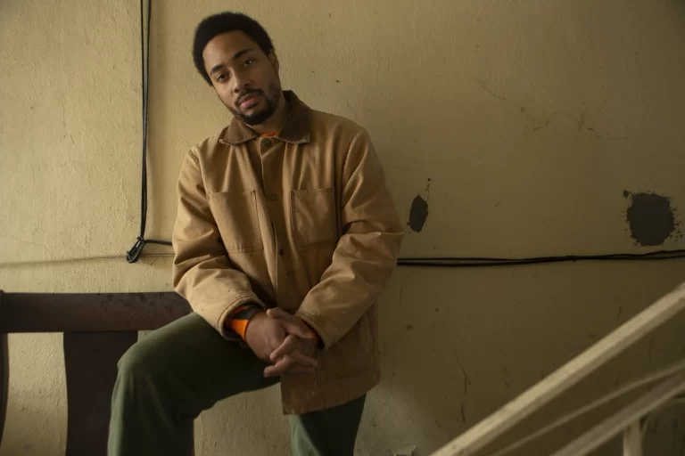 Cadence Weapon posing in front of wall, kneeling on leg, wearing beige jacket