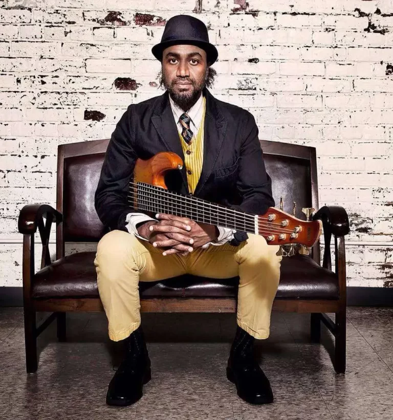 Photo of Waleed seated in front of white brick wall and holding guitar