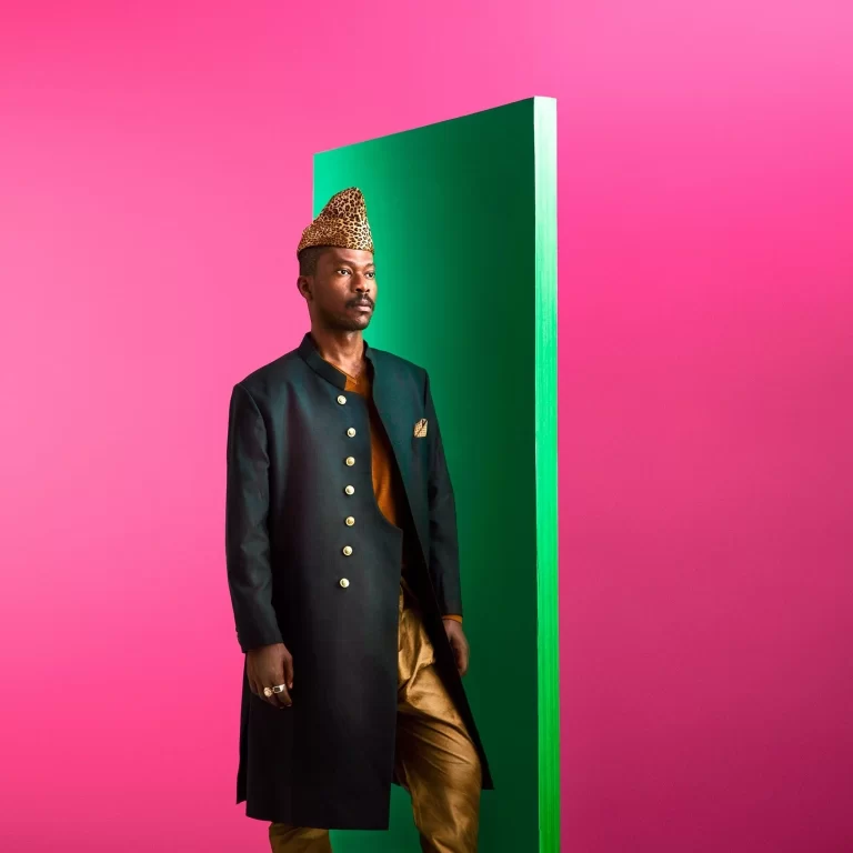 Pierre Kwenders [José Louis Modabi] in front of a bold pink background while wearing a fashionable golden attire, with a short green wall to Pierre's left