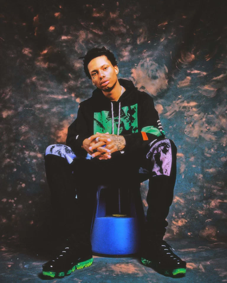 Night Lovell sitting on a blue stool, arms resting on legs, hands together fingers crossed, and his head leaning to his right. The background is a an abstract back and orange backdrop that also covers the floor.