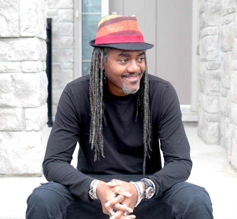 Master T wearing colourful hat, dressed in black looking away, and sitting on the stairs