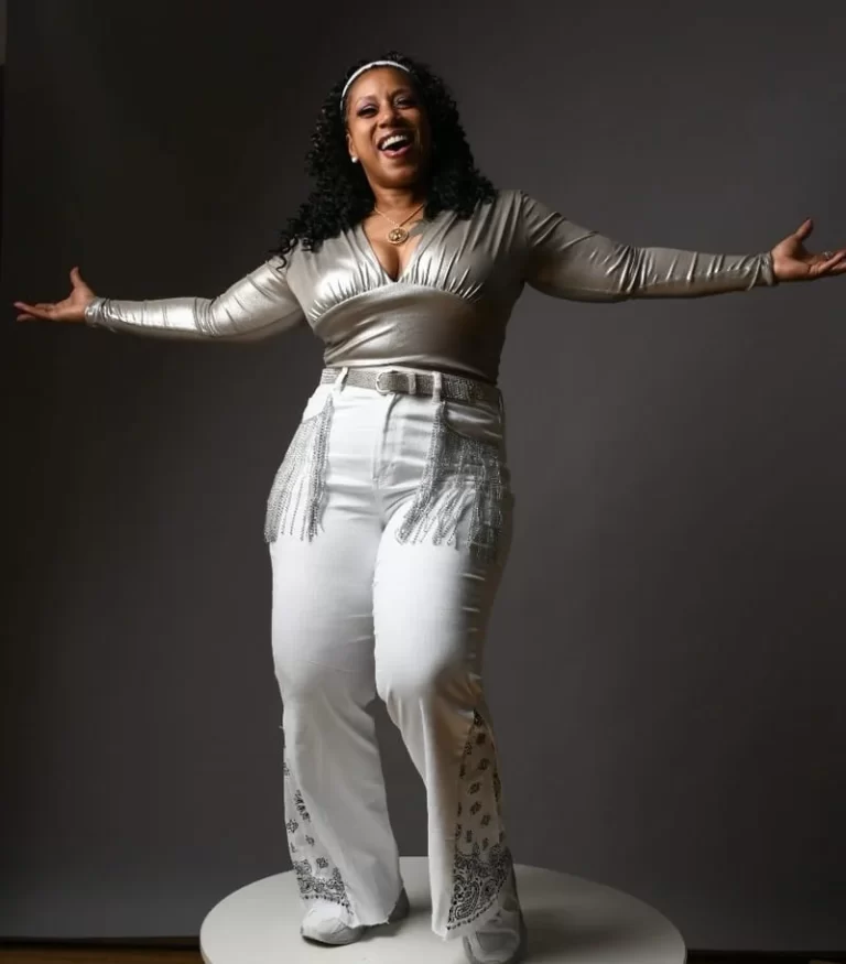 a full-shot photograph of Garnetta Cromwell posing, with her arms spread apart, in front of a grey backdrop.