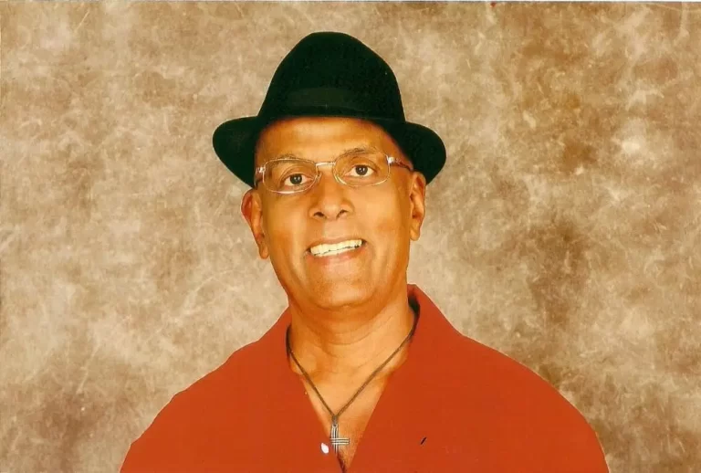 Dick Lochan in red shirt and black hat, posing in front of beige/brown/white background