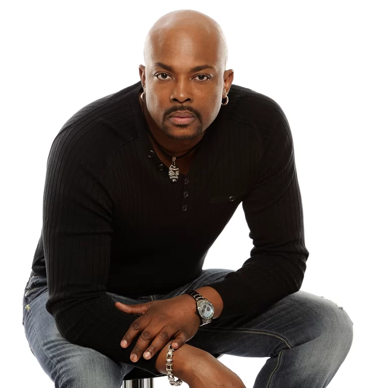 Anslem Douglas crouching in jeans and black shirt, white background