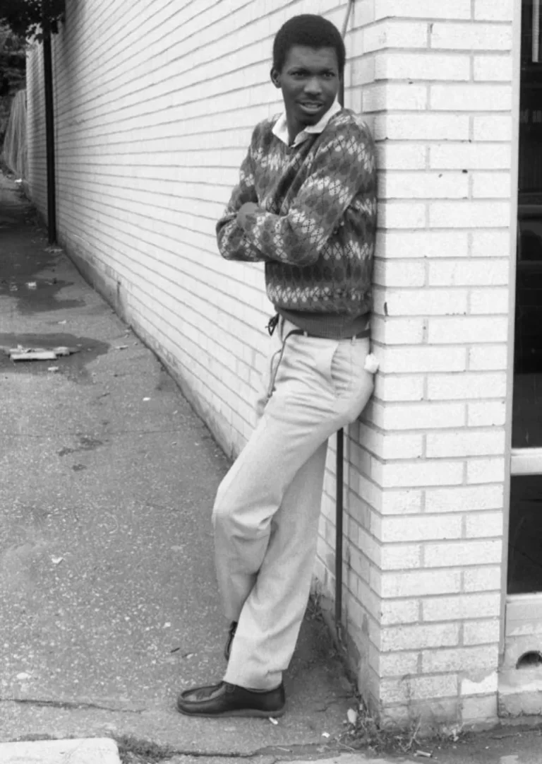 Full-shot of Noel Ellis in a black and white photograph within a daytime exterior. Noel Ellis is seen leaning on a brick wall with his arms crossed.