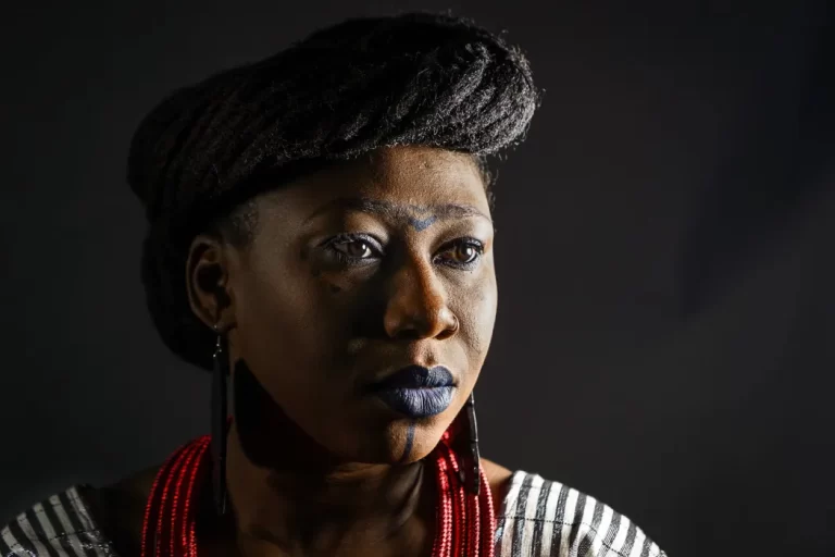 Close-up of Djely Tapa looking to her left, with light being strongly emphasized, all shot in front of a dark grey background