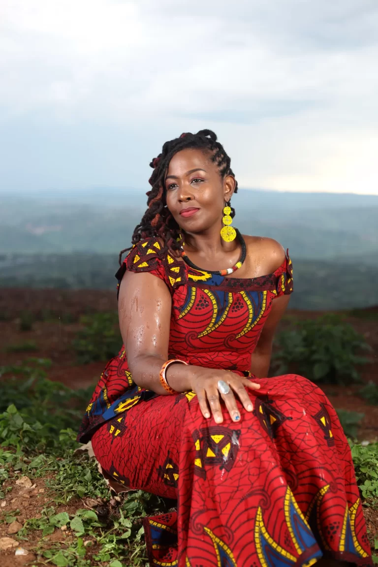 An exterior daytime of a hilled terrain, featuring Empress Nyiringango looking to her right, sitting in a red dress, surrounded by plants and rocks.