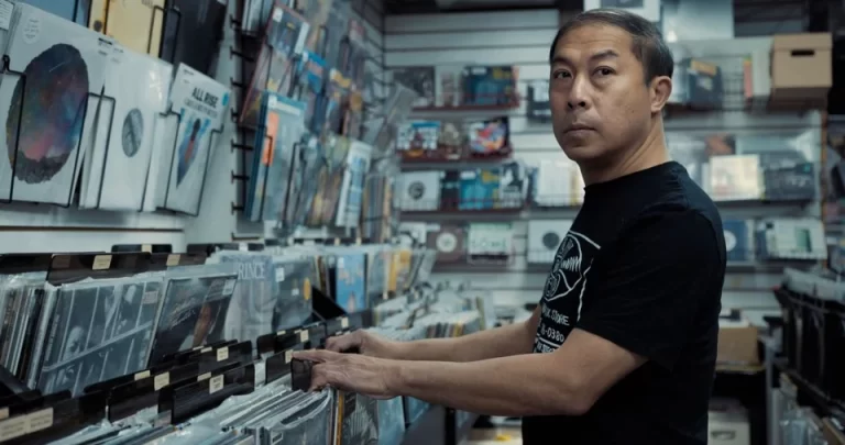 A well-lit interior of a record store, showing a medium shot of Eugene Tam going through records with both hands, while starring to his left.