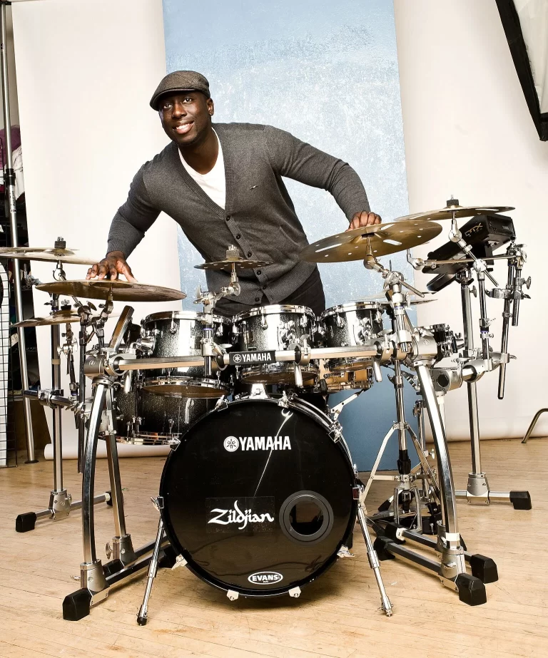 A brightly lit interior of a room with a white backdrop, featuring a full shot of Larnell Lewis standing over a drum kit