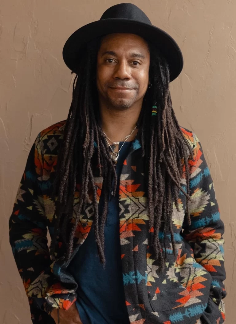 Direct shot of Taylor wearing a black hat and an unbuttoned colourful shirt, smiling