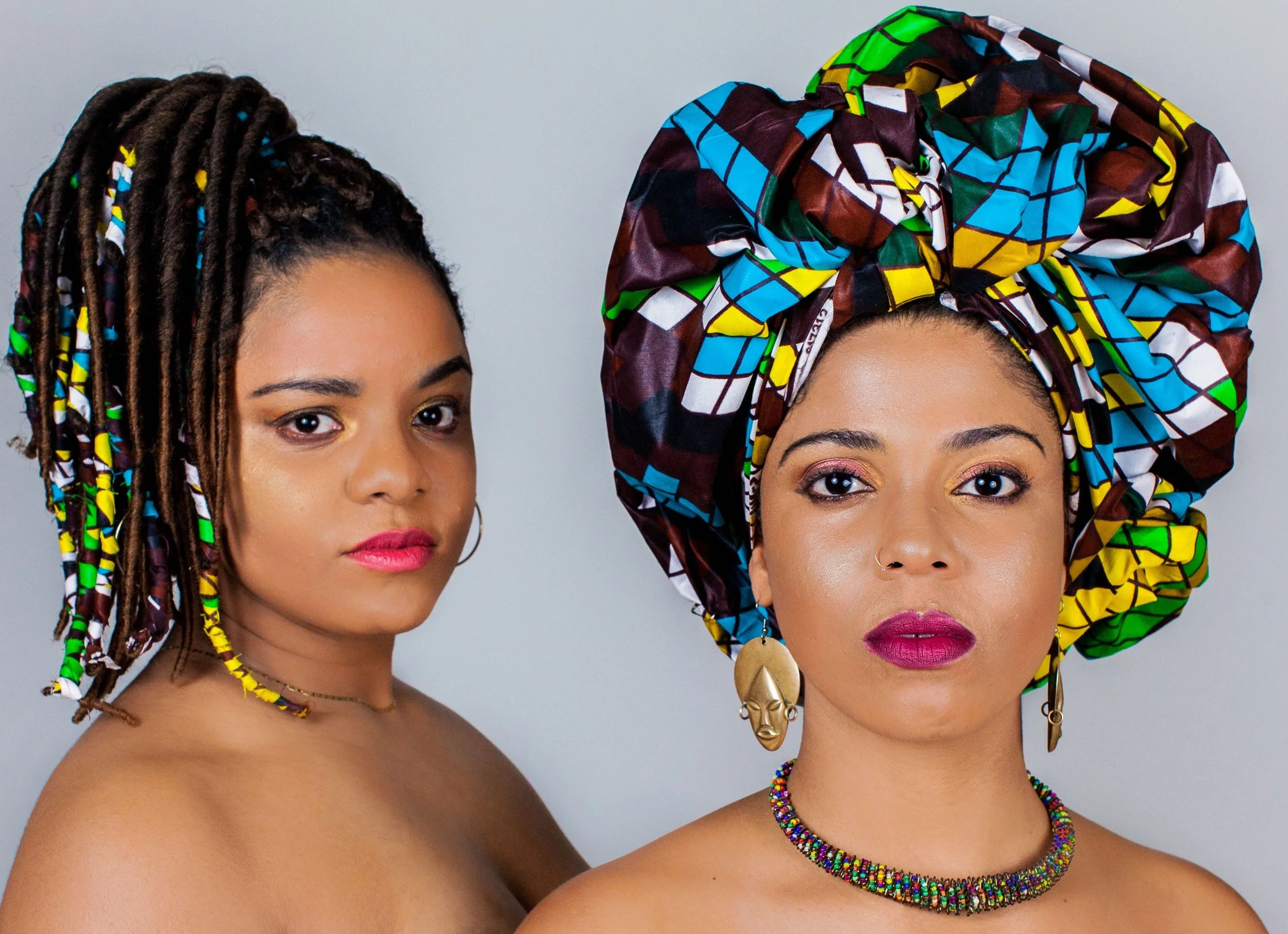 Duo in front of light grey background, naked shoulders, and one wearing a turban/headdress