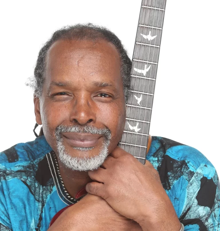 Maurice Gordon holding his guitar in a blue shirt, white background