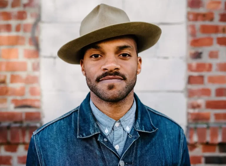 Khari posing with hat, wearing jean jacket, in front of brick wall