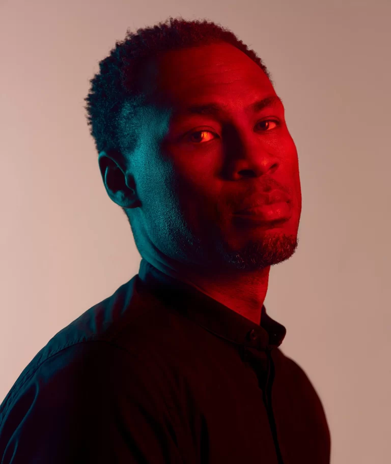 Mark Campbell posing with red lighting on face, beige camera