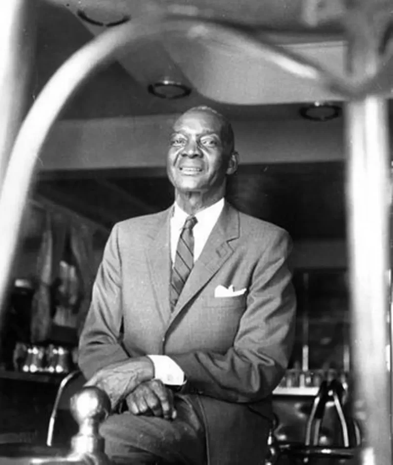 Black and white photo of Rufus Rockhead, seated in bar with legs crossed