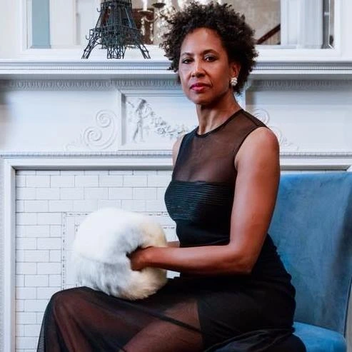 Maria Thompson Corley seated in black dress on blue chair, looking into camera