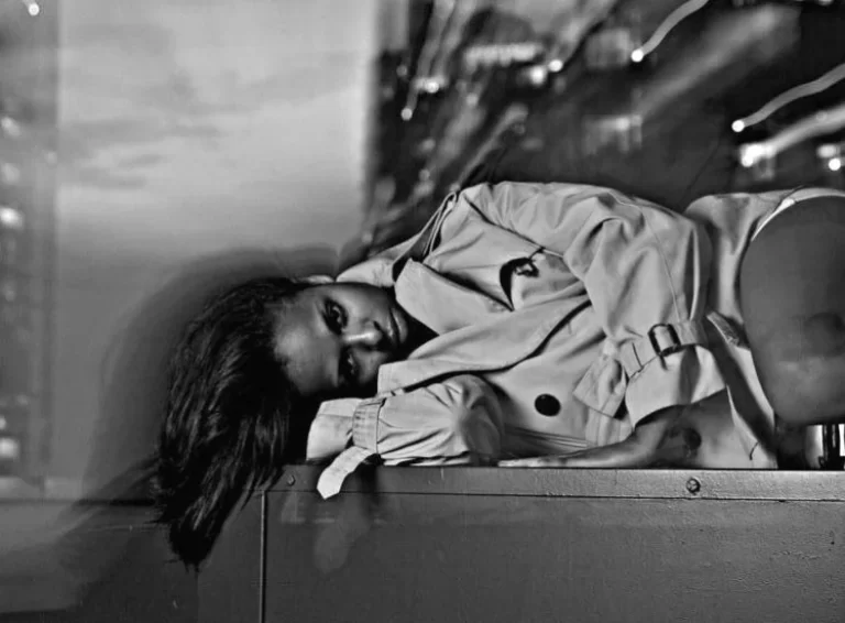 Black and white photo of Graph Nobel lying on her side wearing a trenchcoat, looking into camera