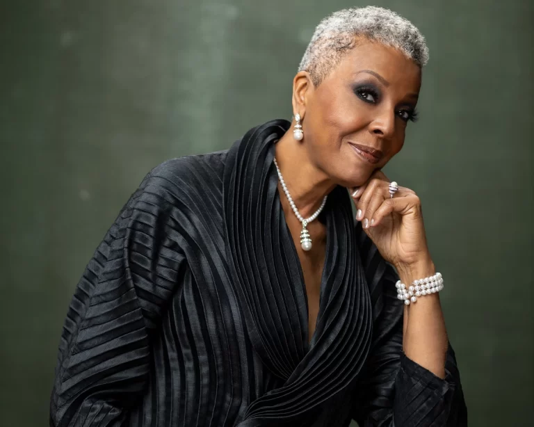 Dee Daniels in front of green background wearing black, hand resting on head which is tilted to the side