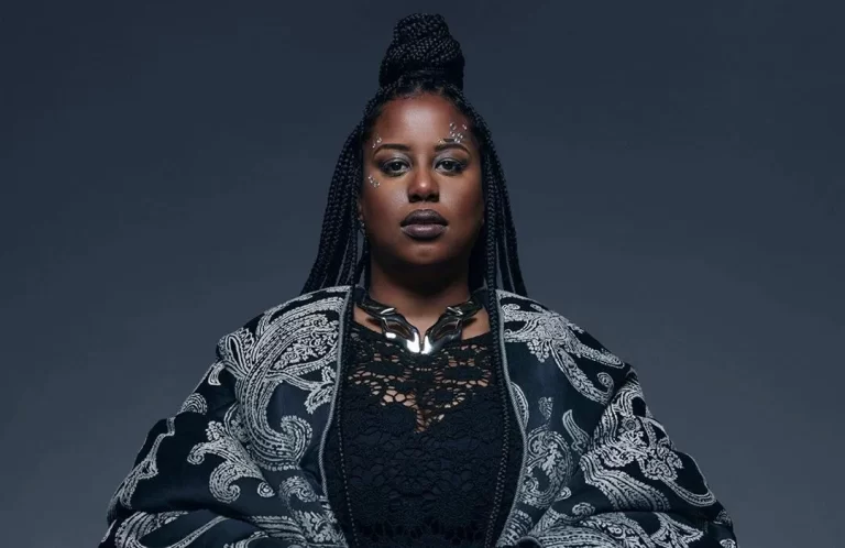 Witch Prophet looking into camera in black and white shawl and chunky black necklace, blue/grey background