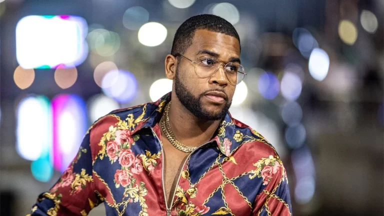 Rochester wearing silk shirt and glasses, looking off camera, blurred lights in background