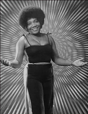 Cynthia Joyce Webber standing in black jump suit, arms outstretched and smiling, white rays effect in background