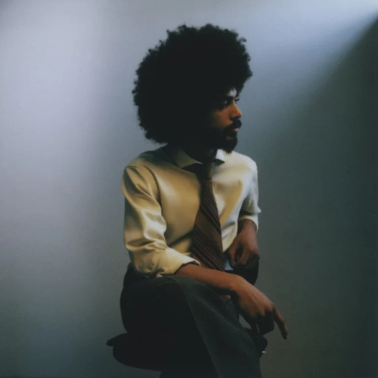 Yves Jarvis in dress shirt and tie, legs crossed and looking to the side